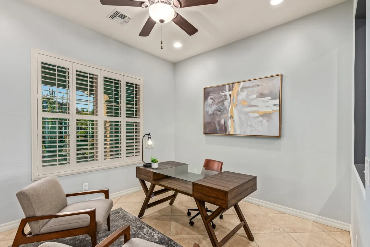 A serene, well-appointed office space with a desk, contemporary artwork, and a comfortable seating area near a window with shutters. The setting is quiet and focused, ideal for productive work or confidential meetings.