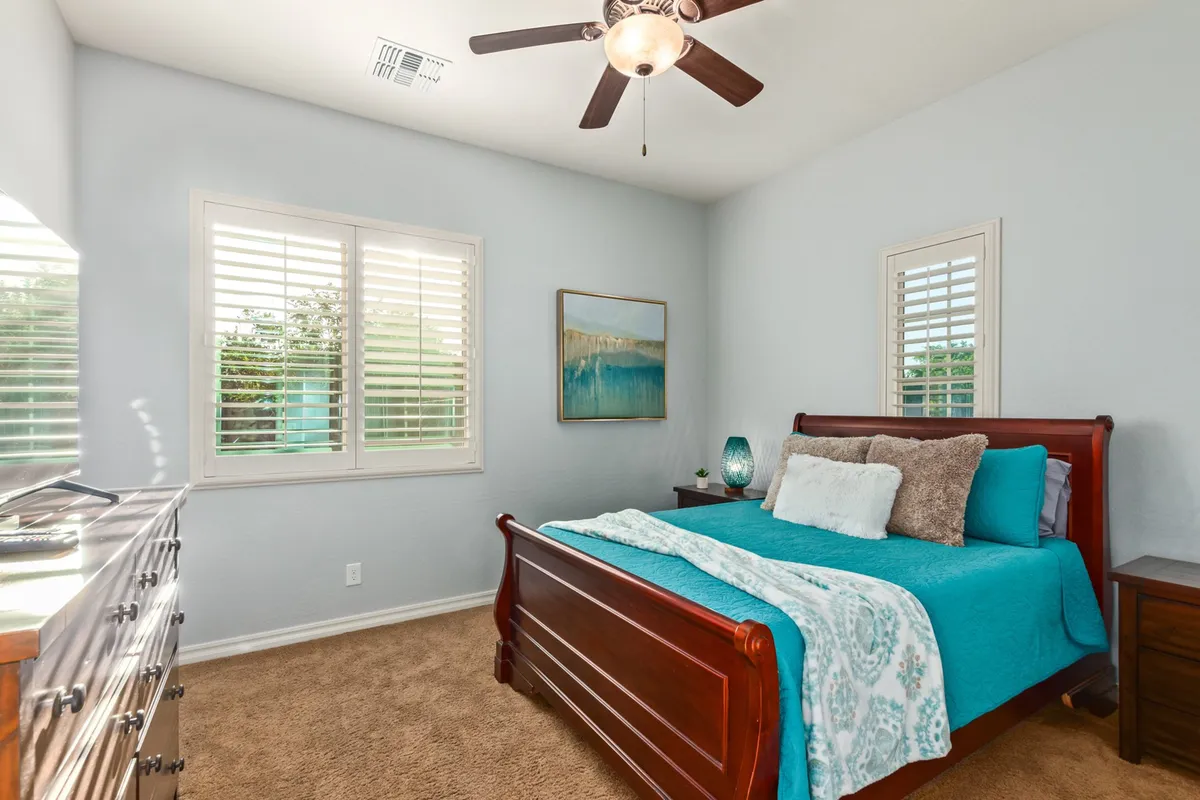 A tranquil bedroom featuring a king bed with turquoise bedding, a wooden dresser, tasteful wall art, and a ceiling fan. Natural light streams in, creating a peaceful, restful atmosphere ideal for relaxation or reading.