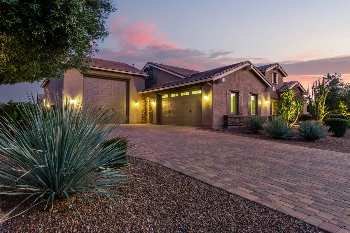 The image features a welcoming exterior view of the home's driveway and front entrance, set against beautifully maintained landscaping and illuminated by the evening light. The secure, gated aspect and spacious setting convey privacy and a sense of arrival.