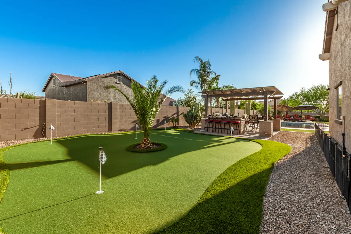 A broad view of the backyard showcasing a private putting green bordered by a palm tree, an outdoor dining area ideal for alfresco meals, and the swimming pool in the background. The setting exudes leisure and activity, with multiple features for play and relaxation.