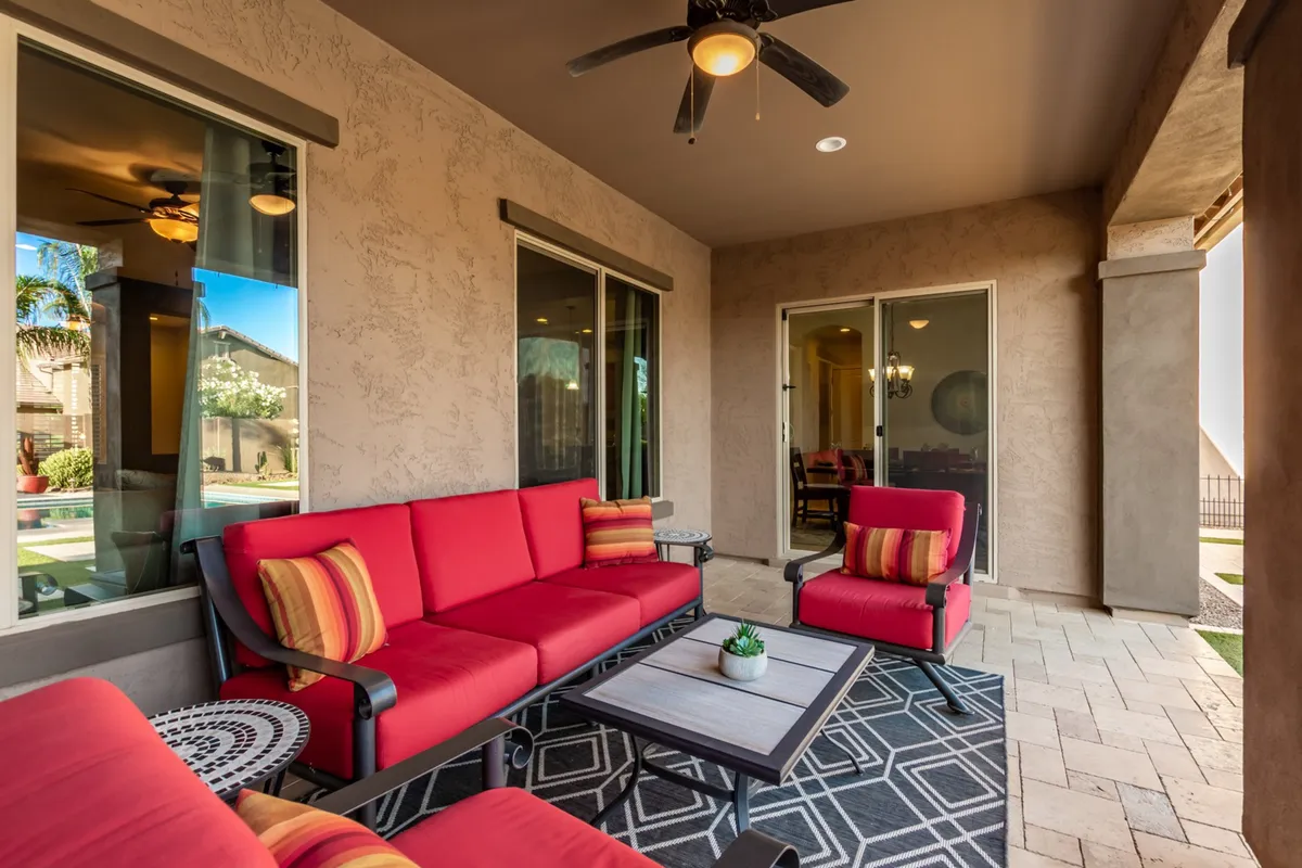 A stylish outdoor patio area with vibrant red seating, a decorative rug, and large glass doors that seamlessly connect the indoor and outdoor living spaces. The setting is shaded, inviting, and ideal for both relaxing alone or hosting guests, while offering views of the garden and sky beyond.