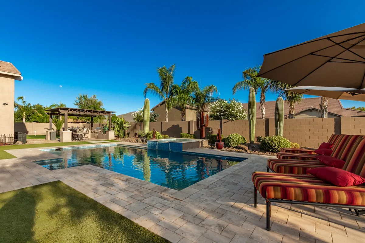 An inviting backyard scene featuring a shimmering swimming pool surrounded by lounge chairs, a stylish pergola for shaded seating, and lush palms and cacti creating a tranquil, resort-like ambiance. The fire pit and ample patio space suggest an ideal setting for both daytime relaxation and starlit gatherings.