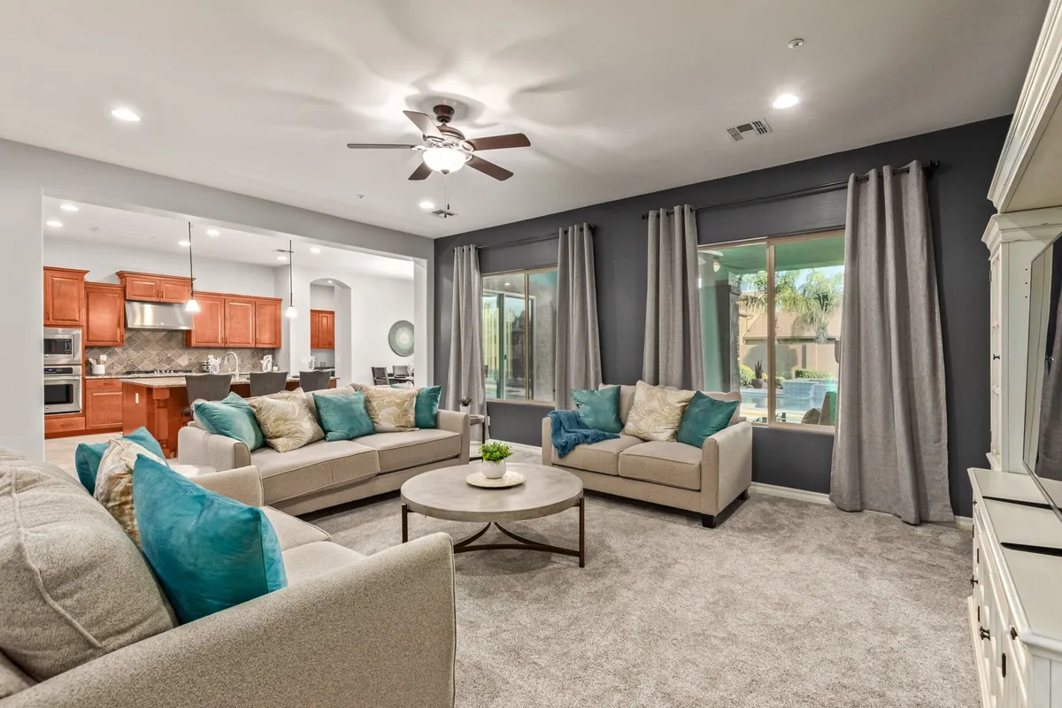 This image presents a spacious and stylish open-plan living area featuring plush sofas with decorative cushions, a round coffee table, and large windows that allow in ample natural light. The kitchen area is visible in the background, seamlessly blending relaxation and dining zones.